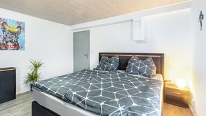 Ein Schlafzimmer mit einem Bett, Nachttischen, einer Wandlampe, einer Deckenleuchte, einer Tür und einer Pflanze.