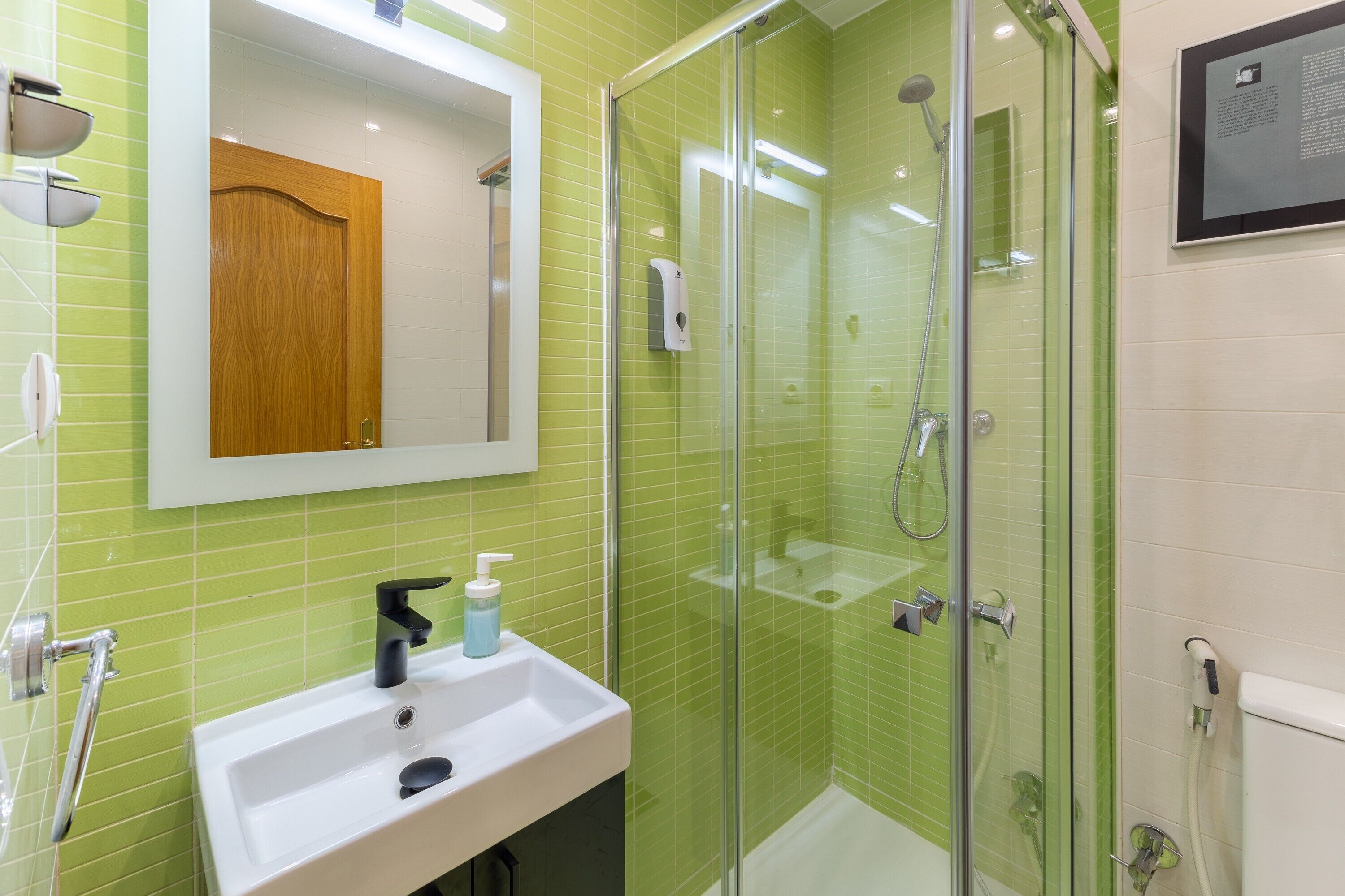 Een badkamer met een douche met glazen wand, een witte wastafel met zeepdispenser en een spiegel boven de wastafel.
