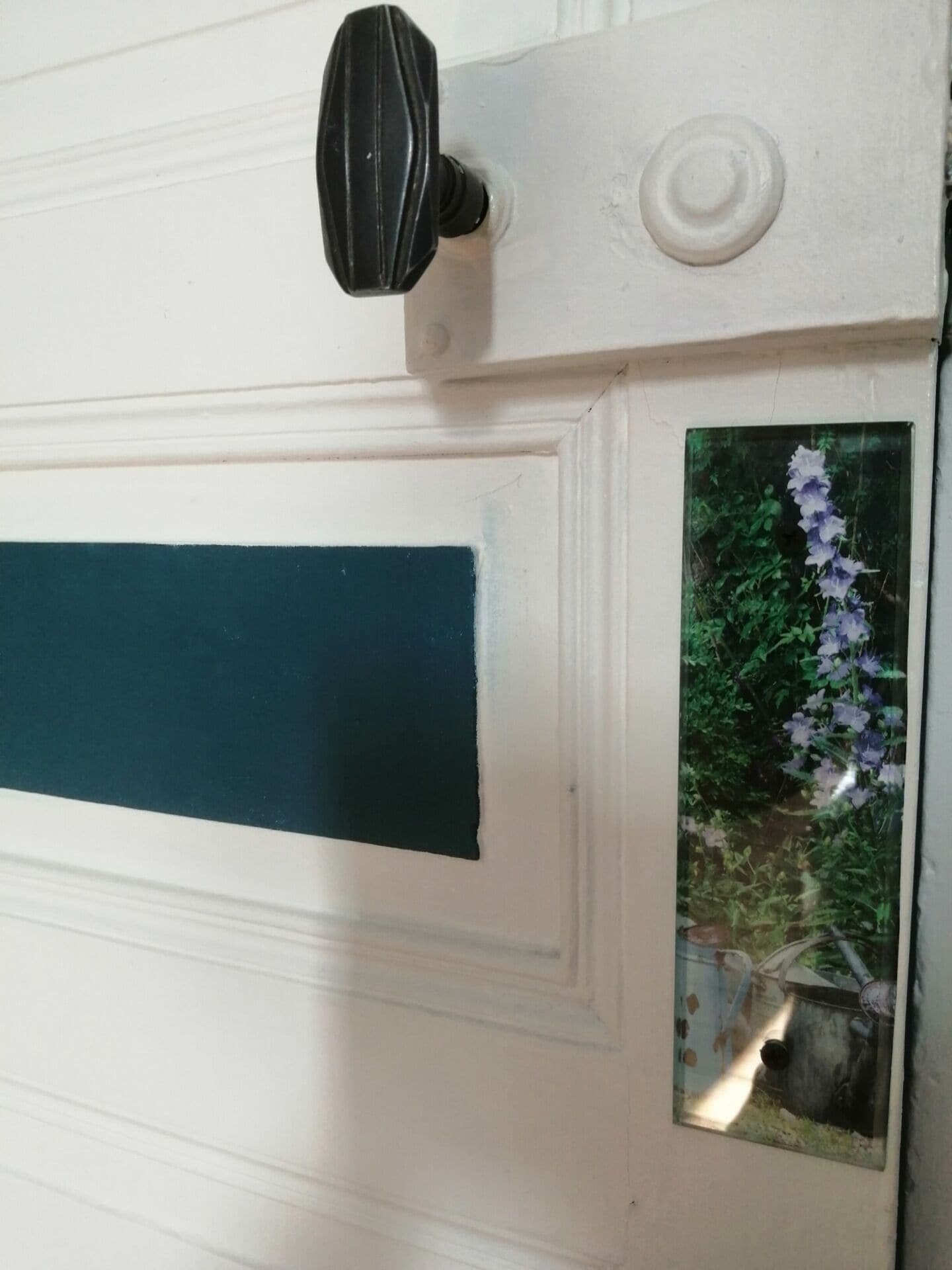 Una puerta blanca con pomo negro, manija verde y un ventanal que muestra flores púrpuras.