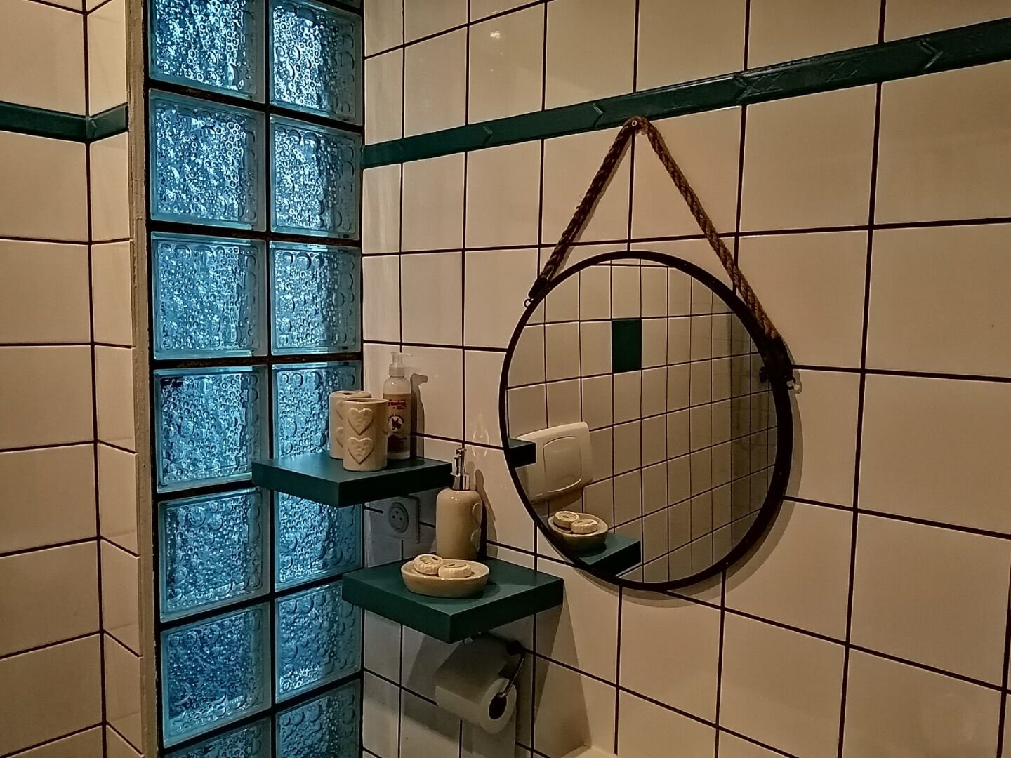 Un baño con pared de azulejos azules, un espejo redondo y estantes con artículos de aseo.