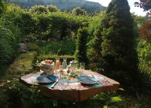 A round wooden table set outdoors with plates, cutlery, a basket of bread, a bottle, and a glass. - La maison O kiwis (Saint-Denis-lès-Martel)