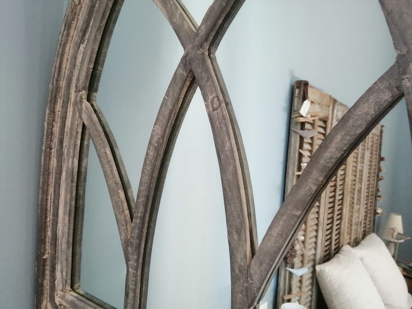 A room with a large, dark wooden mirror with a geometric design, a wicker chair, and a white pillow.