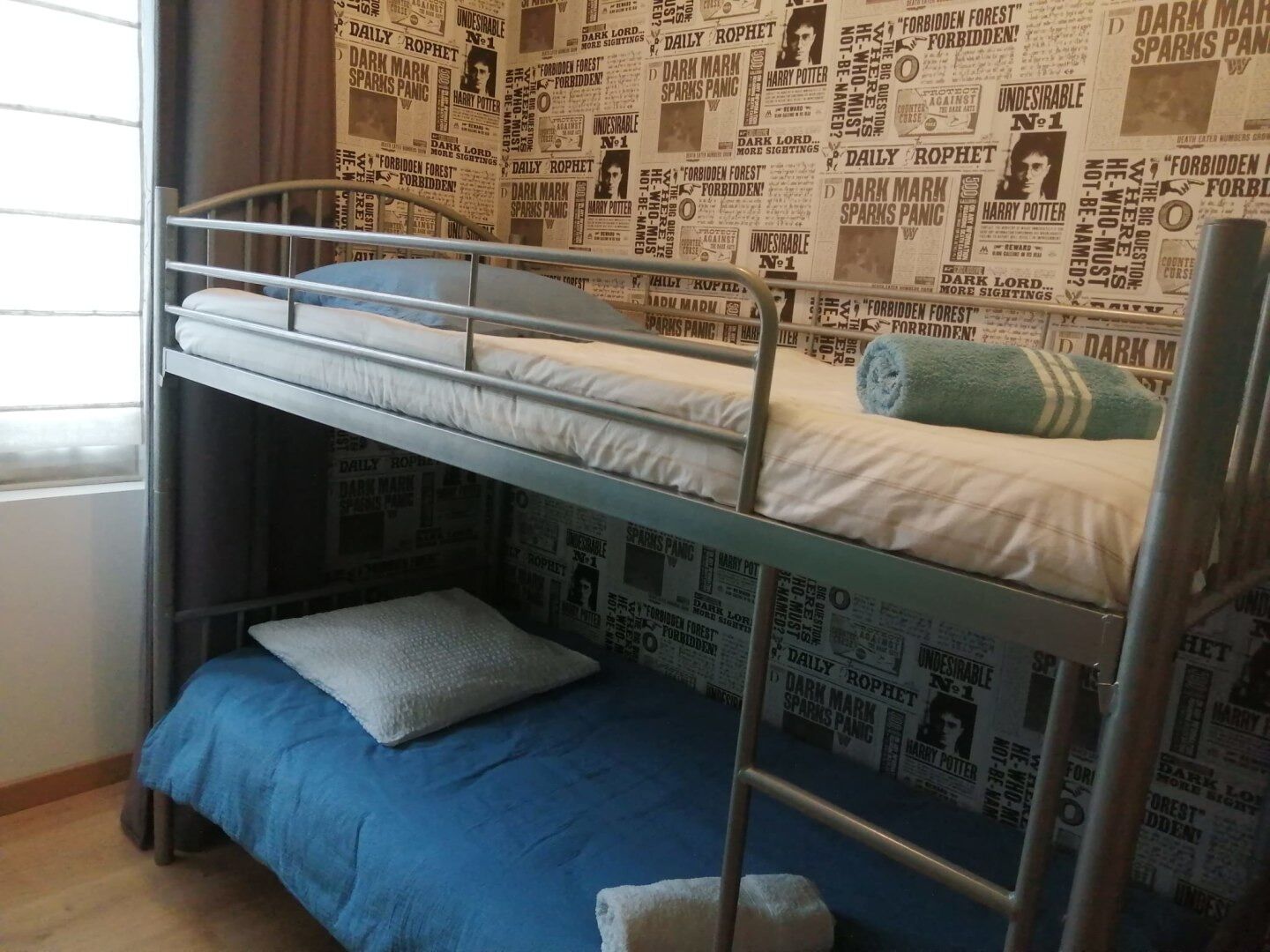 Habitación con literas, un pared cubierta con periódico, una ventana y una colcha azul.