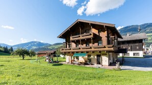 A traditional wooden chalet with a balcony, set in a green field with mountains in the background.