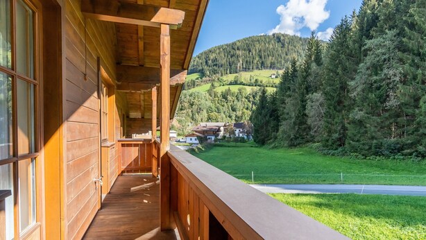 A wooden cabin with a balcony overlooking a green valley and forested hills.