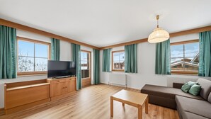 A modern living room with a wooden floor, a flat-screen TV, a sofa, a table, and large windows with curtains.