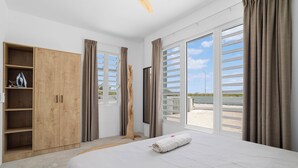 A bedroom with a large bed, wooden wardrobe, and a view of the beach through floor-to-ceiling windows.