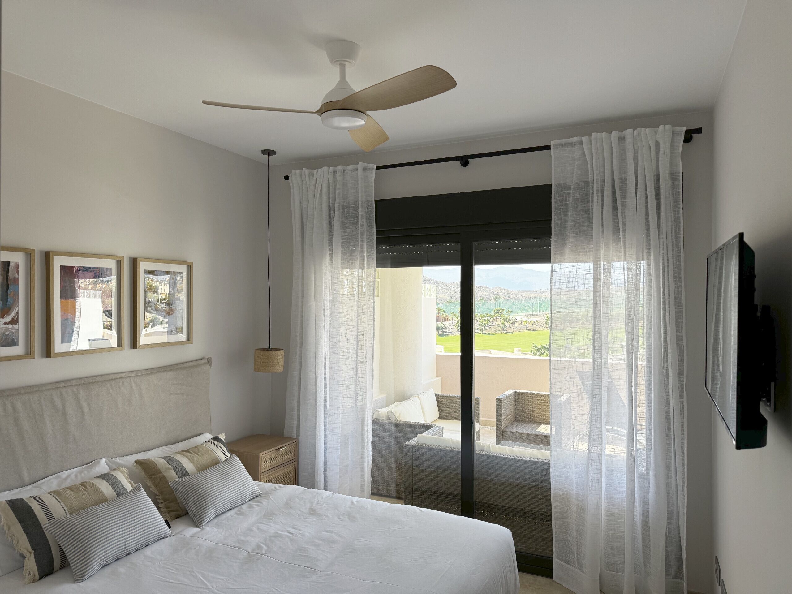 Un dormitorio con cama, ventilador de techo, ventana grande con cortinas translúcidas y vista a un balcón.