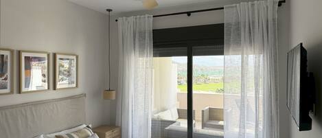 Un dormitorio con cama, ventilador de techo, ventana grande con cortinas translúcidas y vista a un balcón.