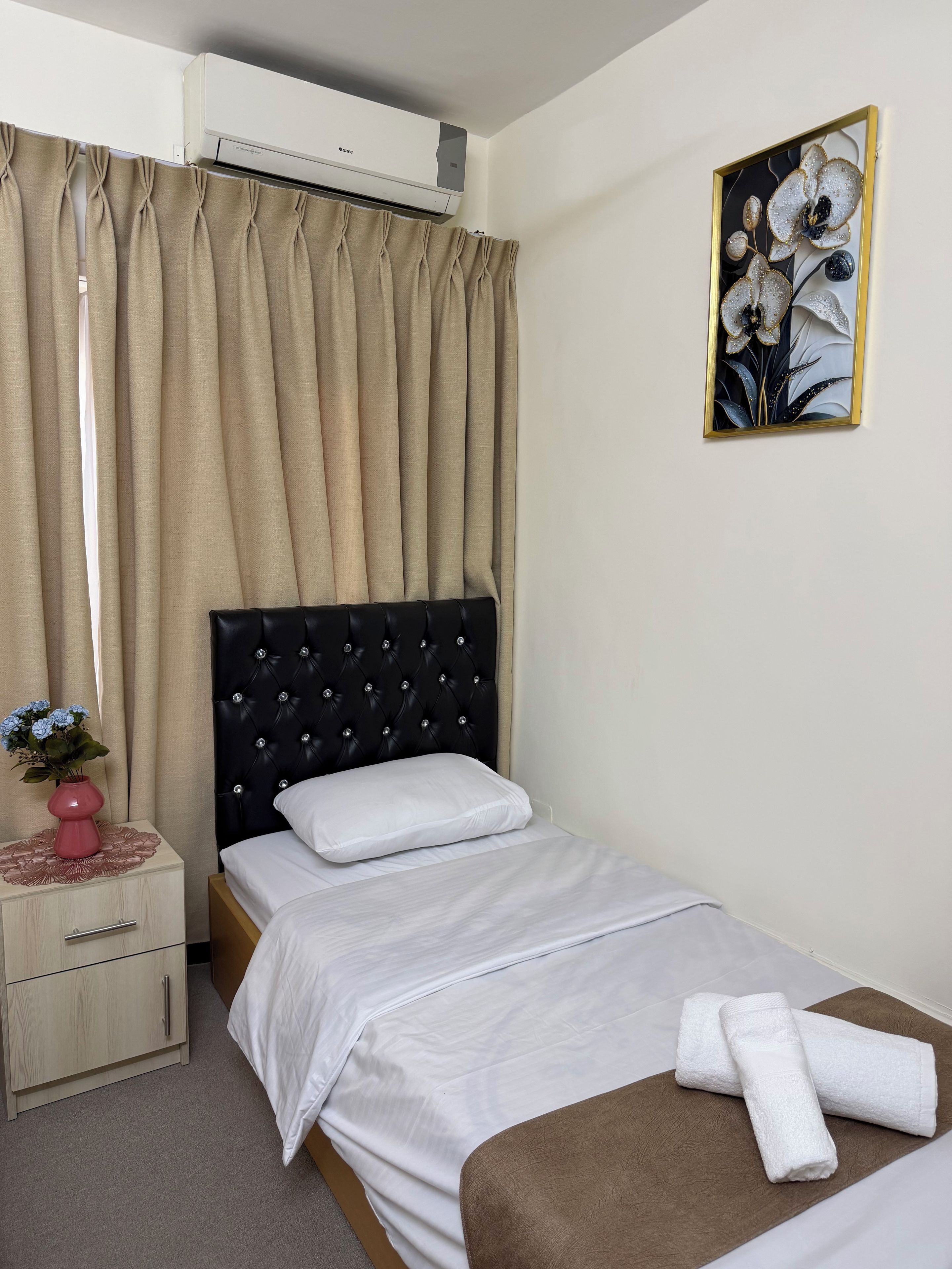 A hotel room with a bed, a nightstand, a drawer unit, an air conditioning unit, and a wall-mounted painting.