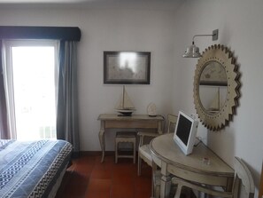 Ein Zimmer mit einem Bett, einem Schreibtisch, einem Sessel, einem kleinen Tisch mit einem Computermonitor, einem dekorativen Spiegel und einem gerahmten Bild an der Wand.