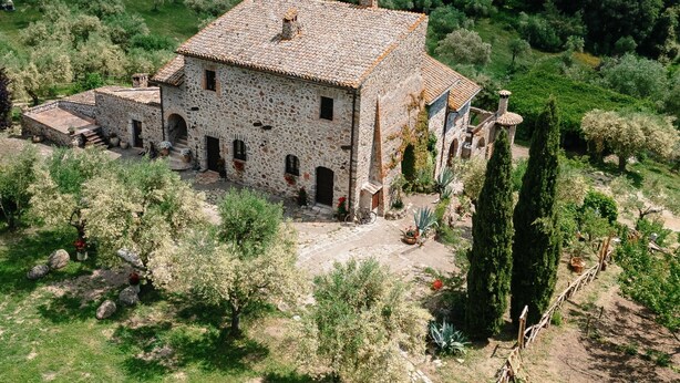 Un bâtiment en pierre, surmonté d’un toit de tuiles, est entouré d’une végétation luxuriante et se dresse sur un arrière-plan de collines verdoyantes.