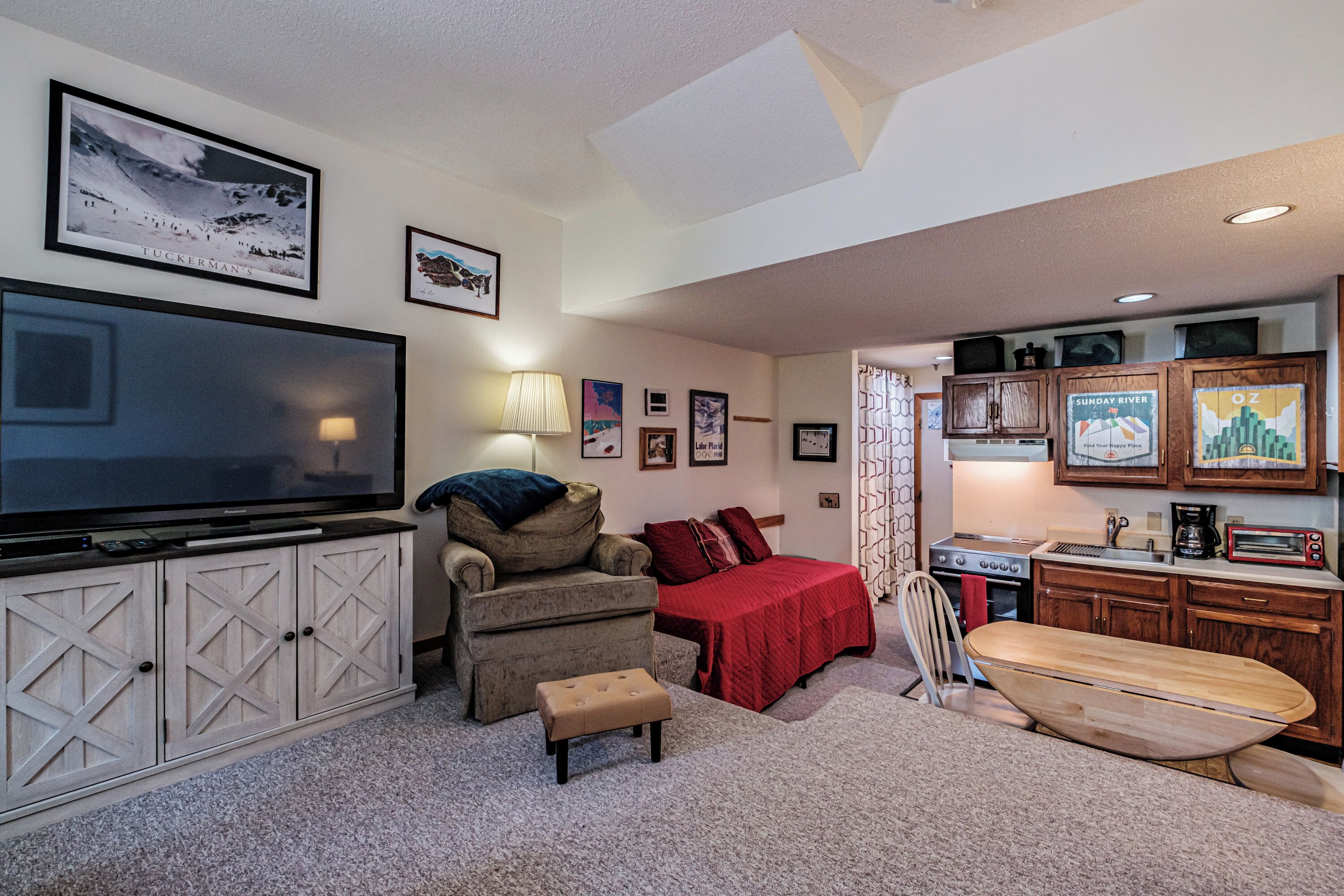 A cozy living room with a large flat-screen TV, a comfortable armchair, a red bedspread, and a wooden surfboard.