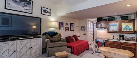 A cozy living room with a large flat-screen TV, a comfortable armchair, a red bedspread, and a wooden surfboard.