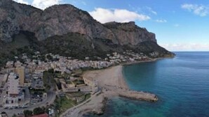 A coastal town with buildings along the shore, a prominent mountain in the background, and clear blue waters. - Villa's apartment on the beach, swimming pool A/C (Palermo)