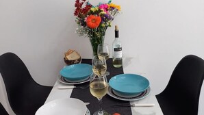 Une table à manger est dressée avec des assiettes bleues, des bols blancs et un verre à vin. Un vase rempli de fleurs colorées et une bouteille de vin sont posés sur la table.
