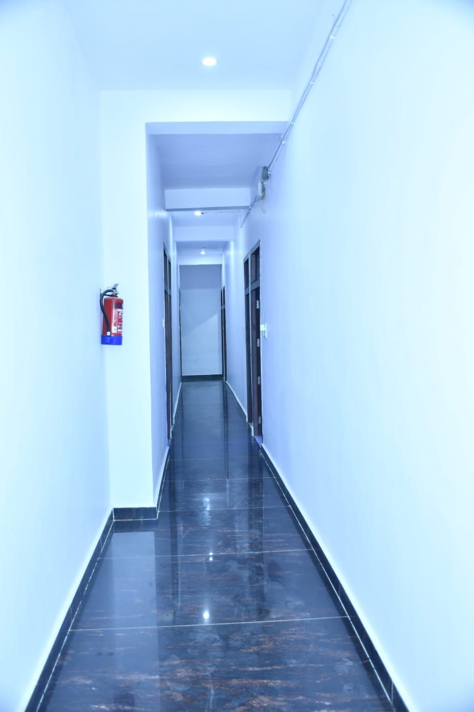 A long, narrow hallway with glossy black flooring and white walls. A fire extinguisher is mounted on the wall to the left.