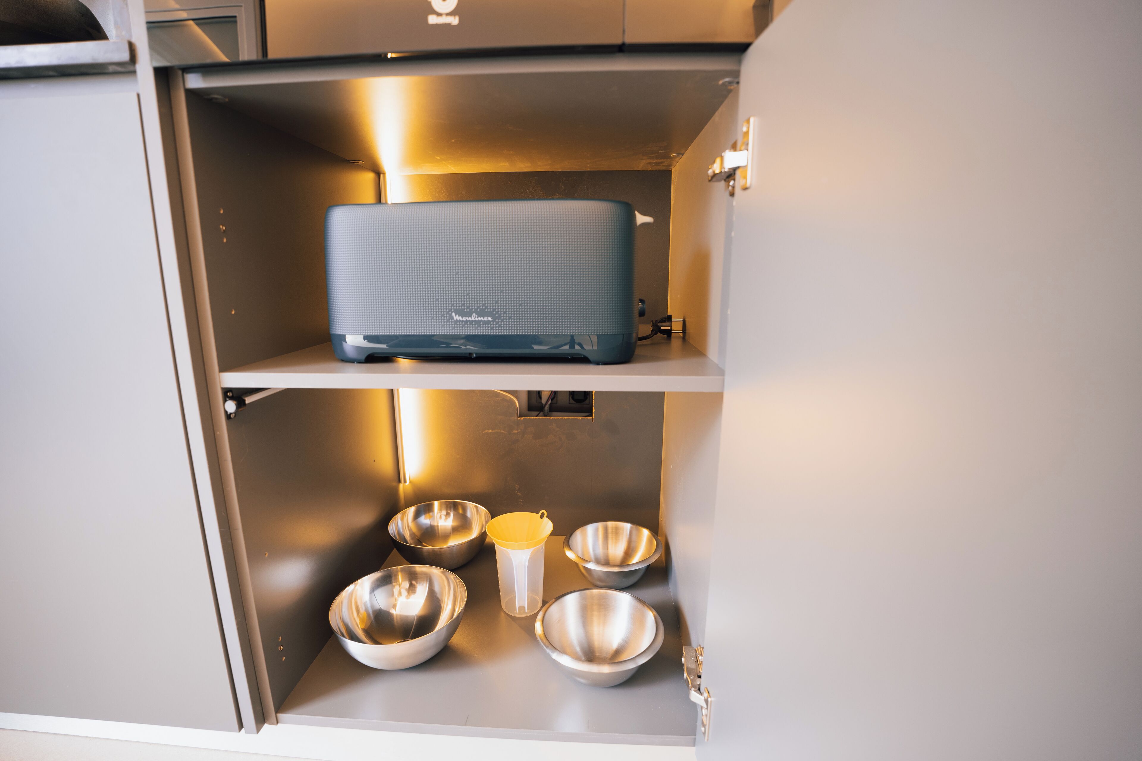 A compact kitchen with a built-in microwave and a shelf above it containing a kettle and four stainless steel bowls.