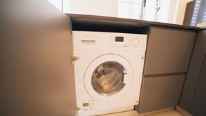 Une petite machine à laver blanche à chargement frontal, installée dans un petit espace buanderie avec un meuble gris et une fenêtre au-dessus.
