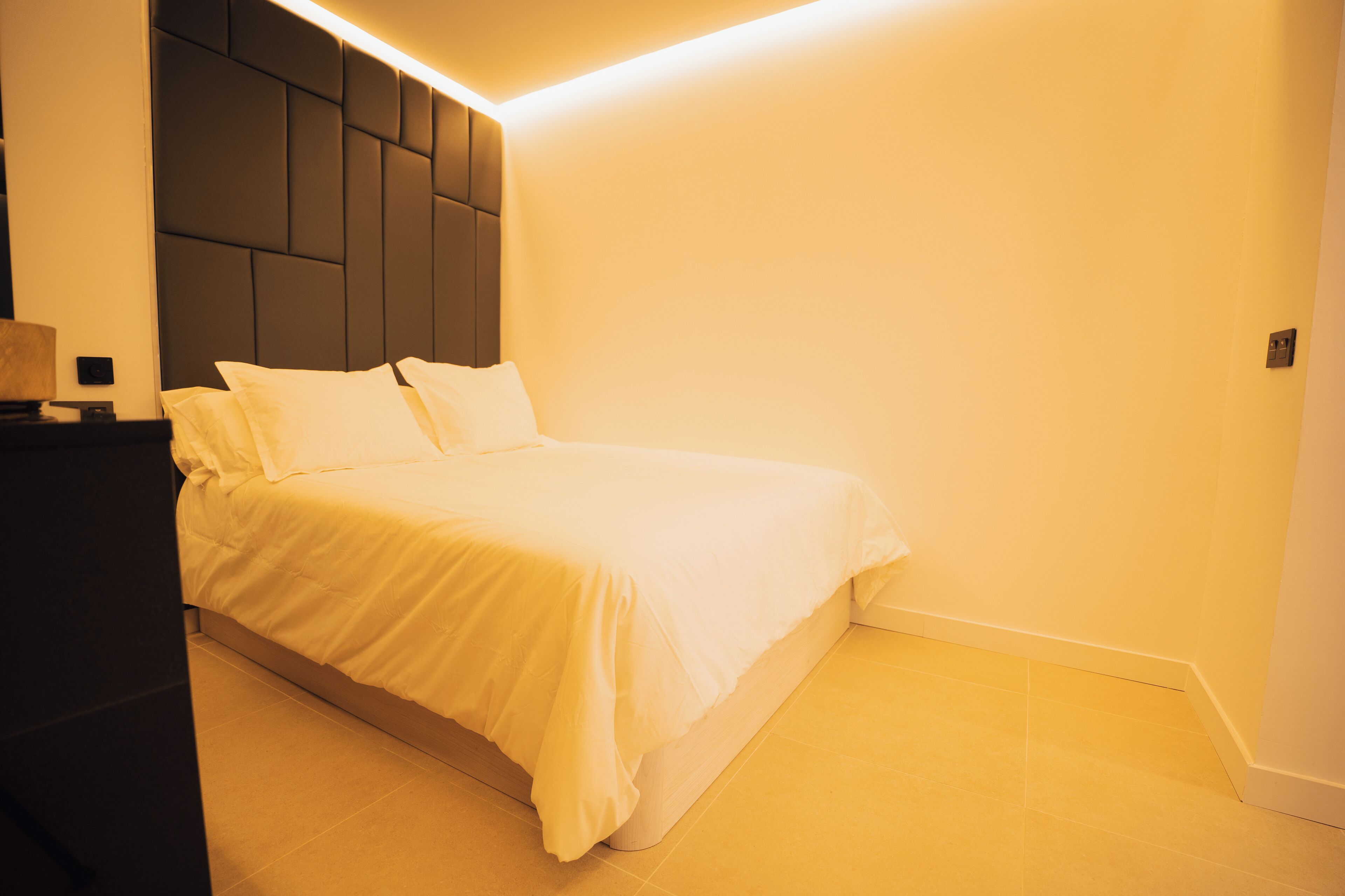 A neatly made bed with white bedding in a room with a modern design, featuring a dark headboard and light-colored walls and floor.