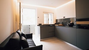 Une cuisine moderne avec des meubles gris, un plan de travail noir et un canapé noir dans le salon attenant.