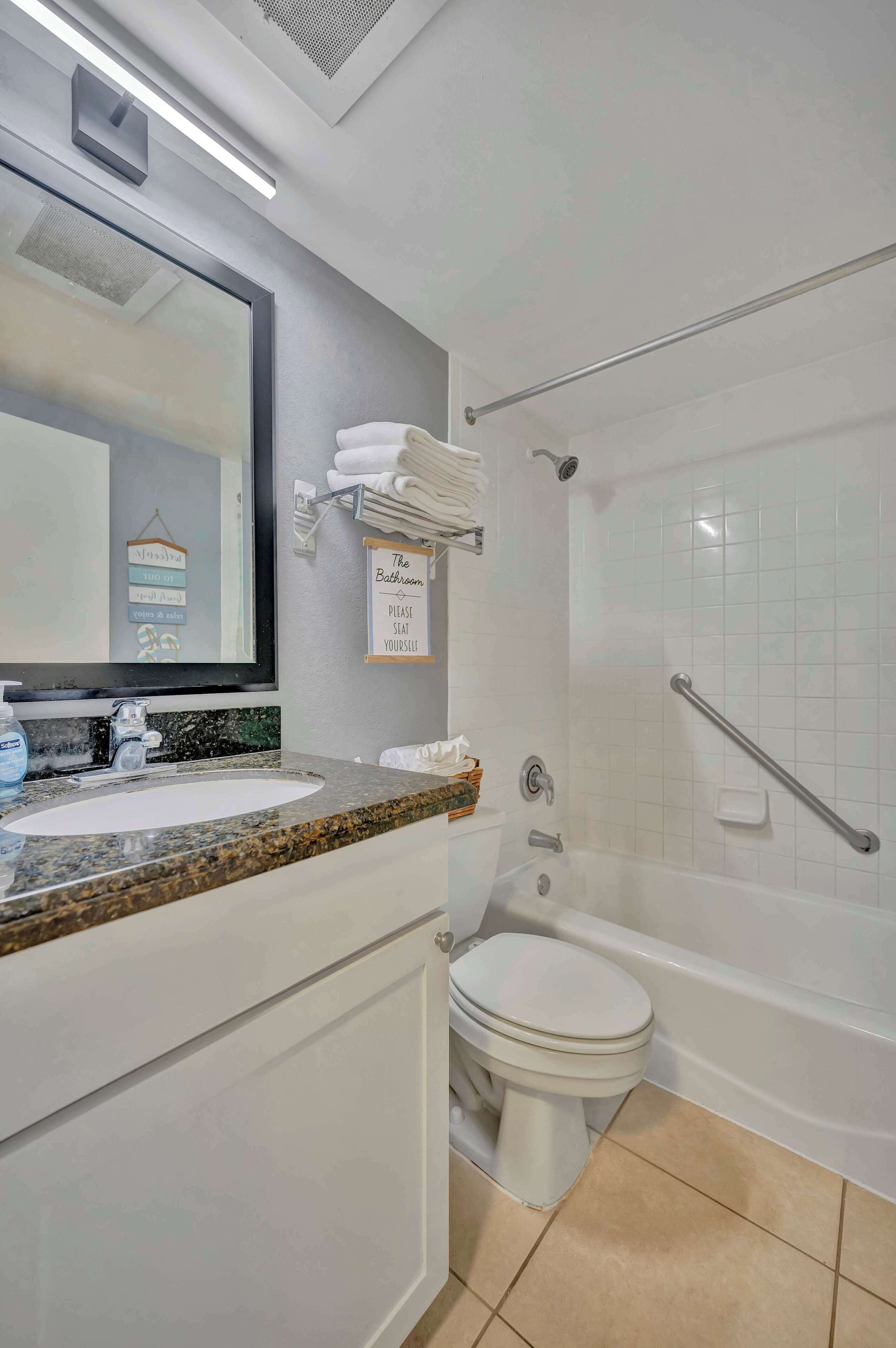 A modern bathroom with a granite countertop, a white toilet, a bathtub with a grab bar, and a towel rack with folded towels.