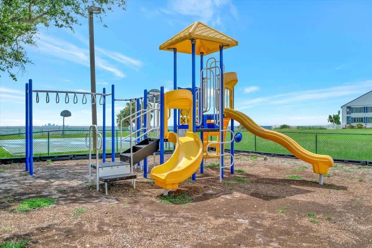 Un parque infantil con un tobogán amarillo y estructuras para escalar, rodeado por una cerca y un edificio al fondo.