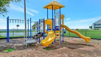 A playground with a yellow slide and climbing structures, surrounded by a fence and a building in the background.
