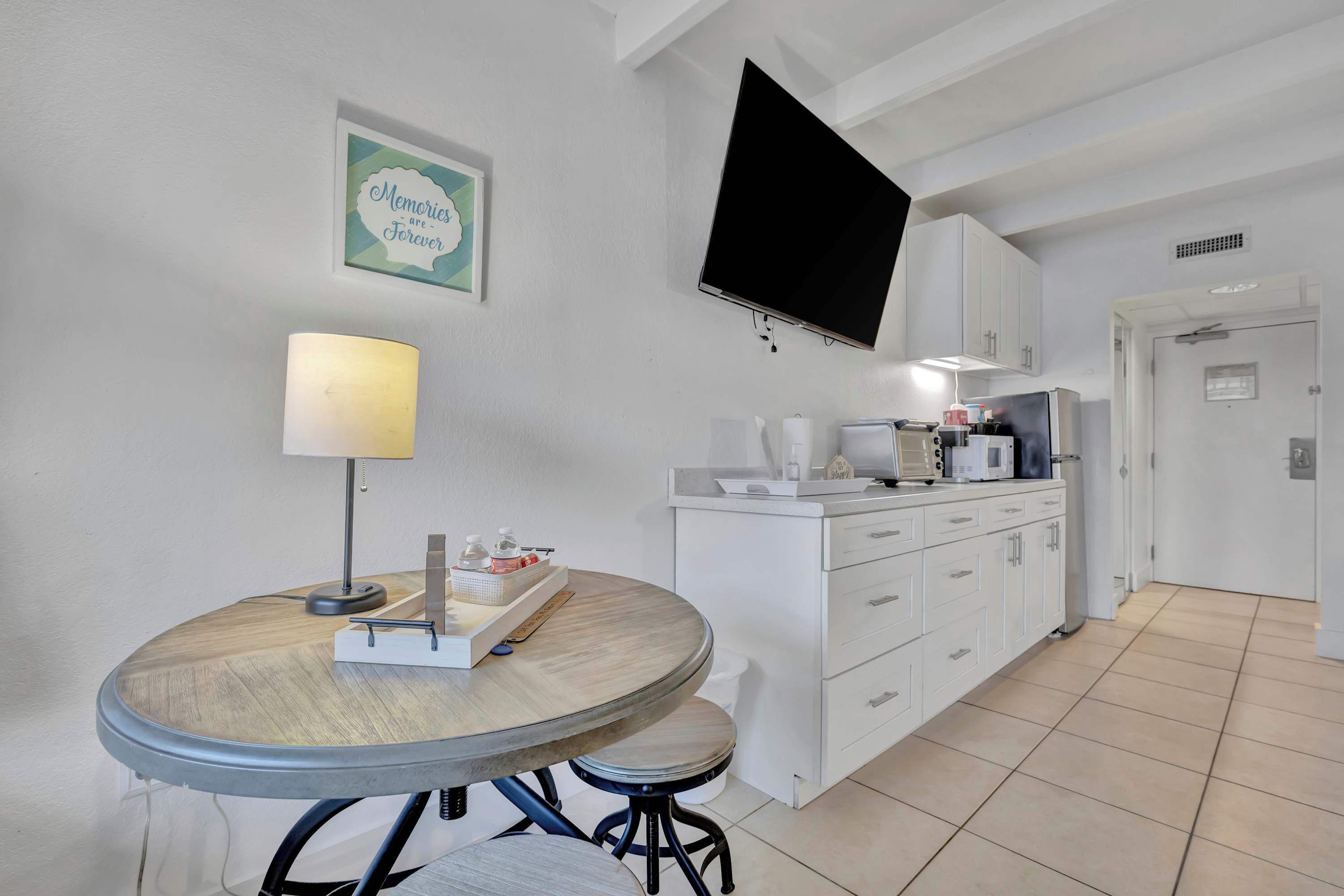Een moderne keuken met witte kasten, een eettafel met een lamp en een flatscreen televisie aan de muur.