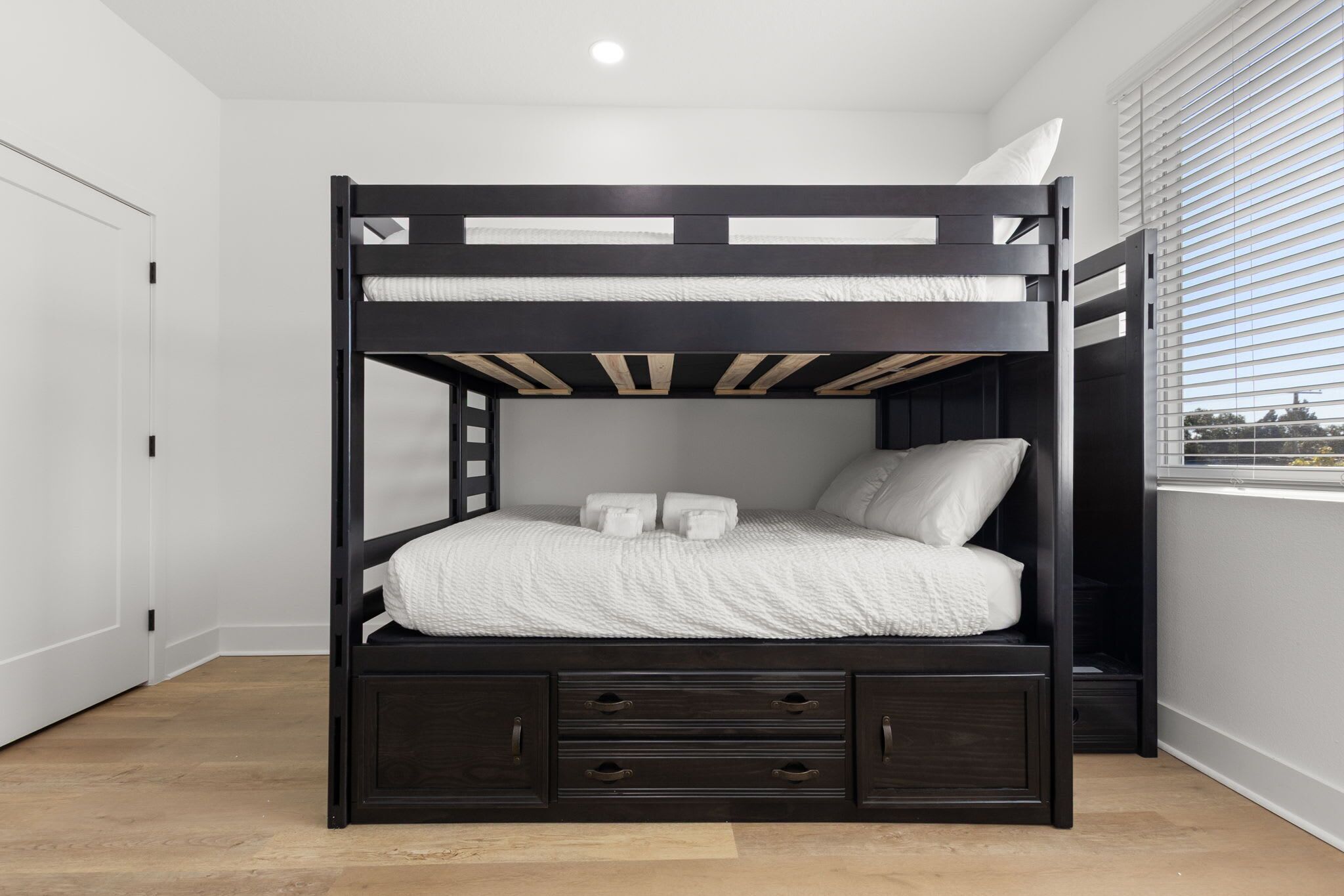 A bunk bed with a white mattress and pillows, a dark wooden frame, and a window with blinds.