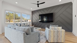 A modern living room with a grey sectional sofa, a patterned ottoman, and a flat-screen TV mounted on a grey wall with a chevron pattern. - NEW Casa Cielo - Gorgeous Newly Built Tampa Home (Tampa)