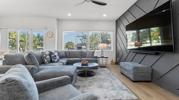 Un salon moderne avec un canapé d’angle gris, une table basse ronde et un pouf gris. Un téléviseur à écran plat est fixé au mur, et un ventilateur de plafond est installé au-dessus.