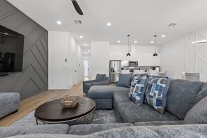 A modern living room with a grey sectional sofa, a wooden coffee table, and a flat-screen TV mounted on the wall.