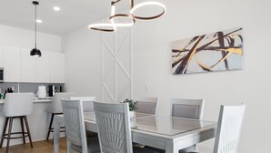 Une cuisine moderne avec des meubles blancs, un coin repas doté d’une table en verre et de chaises à revêtement gris, et une peinture abstraite contemporaine accrochée au mur.