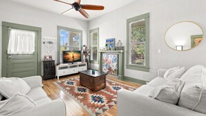 A living room with a ceiling fan, a fireplace, a television, and a large window with blinds. - NEW Lakeland Landing Cute Bungalow by Downtown (Lakeland)