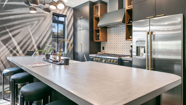 Une cuisine moderne dotée d’un grand îlot central, de meubles de rangement aux couleurs sombres, d’appareils électroménagers en acier inoxydable et d’un luminaire original.
