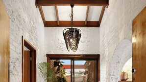 Un pasillo con paredes de ladrillo blanco, vigas de madera, una planta grande en maceta y un ventanal corredera que da a un espacio exterior.