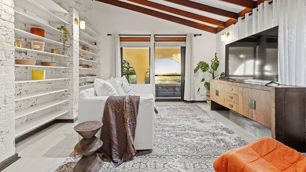 Una sala de estar moderna con pared de ladrillo blanco, vigas de madera, un sofá, un televisor y una alfombra estampada.