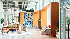 A spacious lobby with a modern design, featuring a central seating area with sofas and chairs, a large abstract painting on the wall, and a staircase in the background.