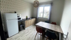 A compact kitchen with a refrigerator, microwave, and wooden cabinets. There is a dining table with chairs and a window with curtains. - Chill & relax (Limoges)