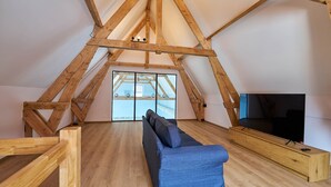 Habitación en el ático con techo de madera, un sofá, un televisor y un ventanal grande.