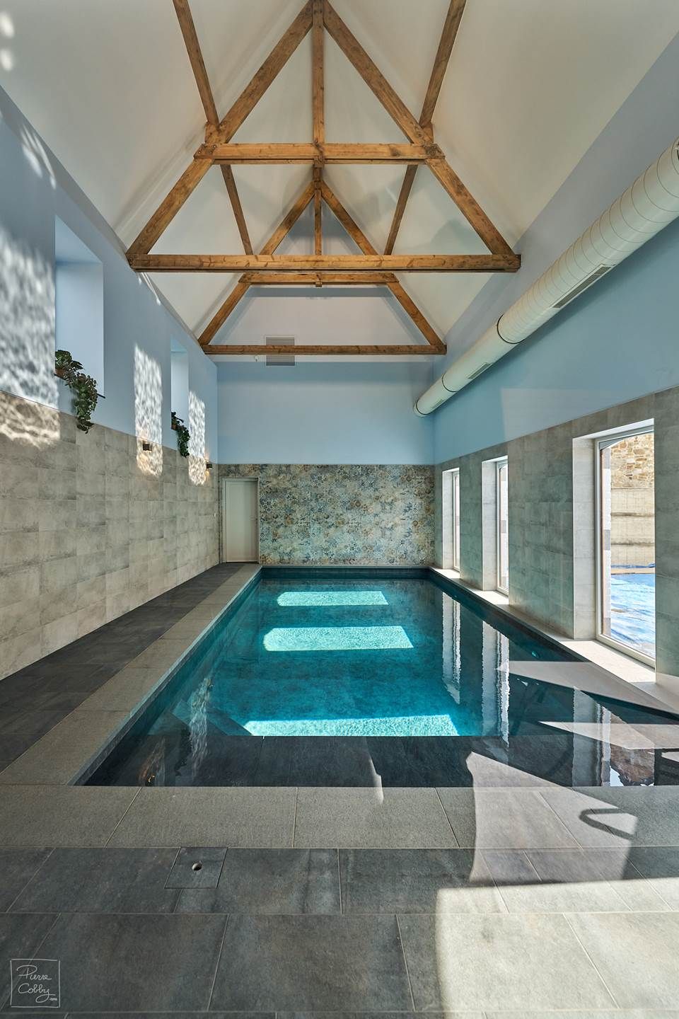 Une piscine intérieure avec de grandes fenêtres offrant une vue dégagée sur le ciel, un mur en pierre et une structure de plafond en bois.