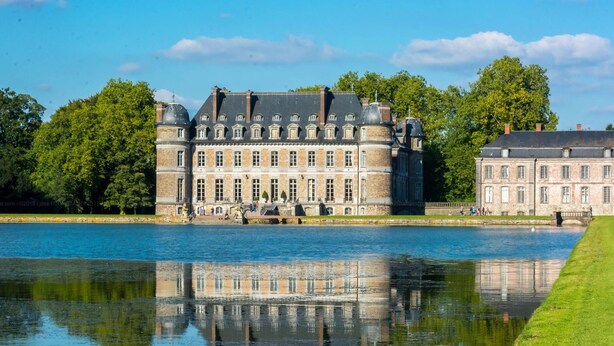 Un edificio grande e histórico de fachada simétrica, con ventanas amplias y una torre central, ubicado junto a una masa de agua tranquila que refleja el edificio.