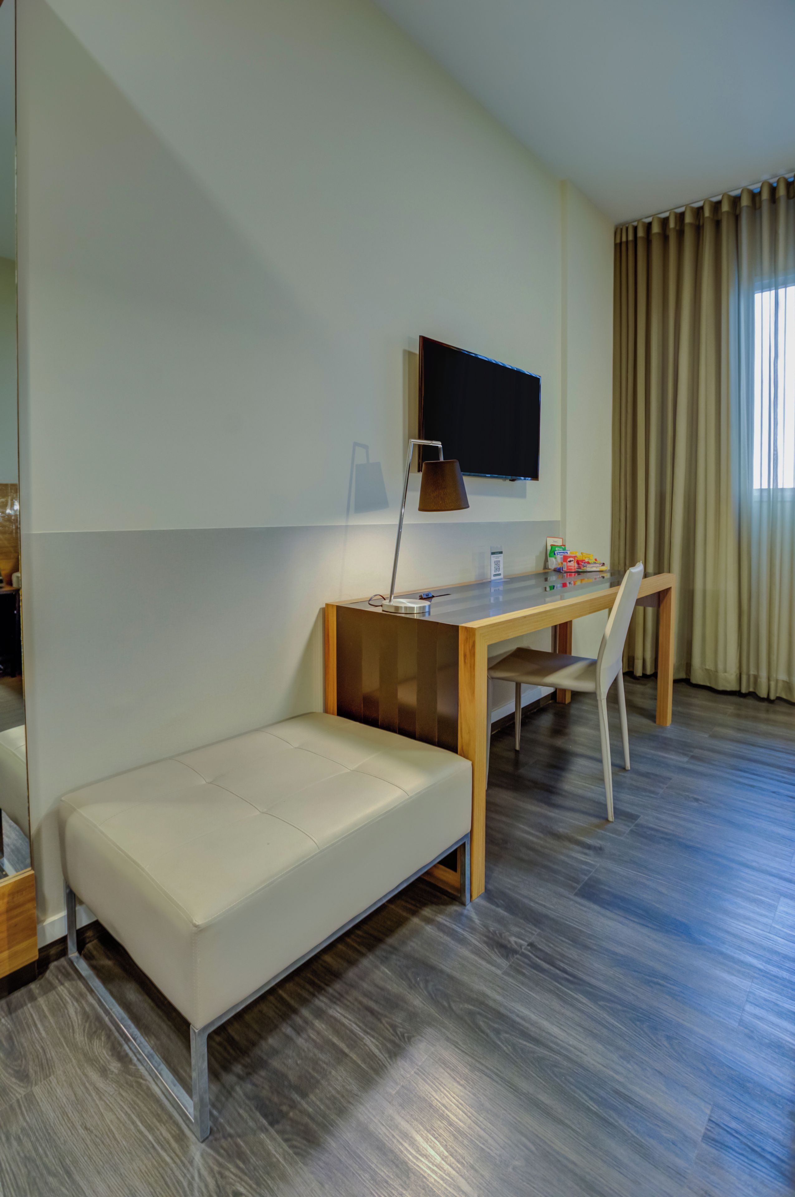 Quarto de hotel moderno com sofá, escrivaninha de madeira, televisão e janela com cortinas.