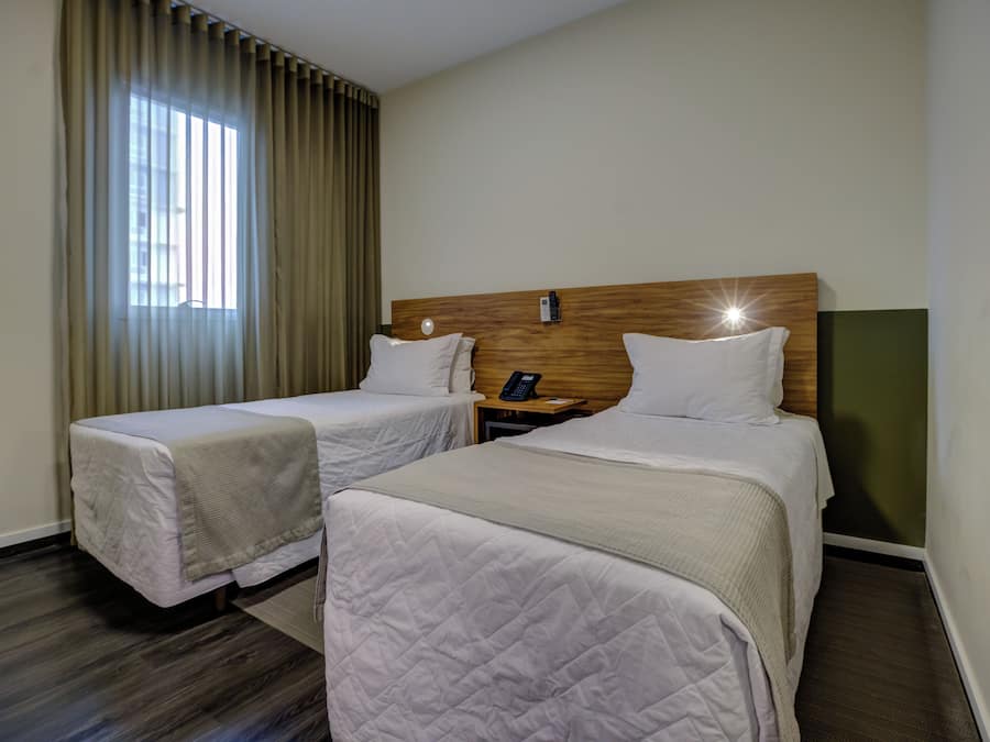Habitación de hotel con dos camas, cabecera de madera, mesita de noche con teléfono y ventana con cortinas.
