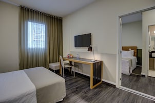 Una habitación de hotel con cama, escritorio con silla, televisor y puerta que da a otra habitación.