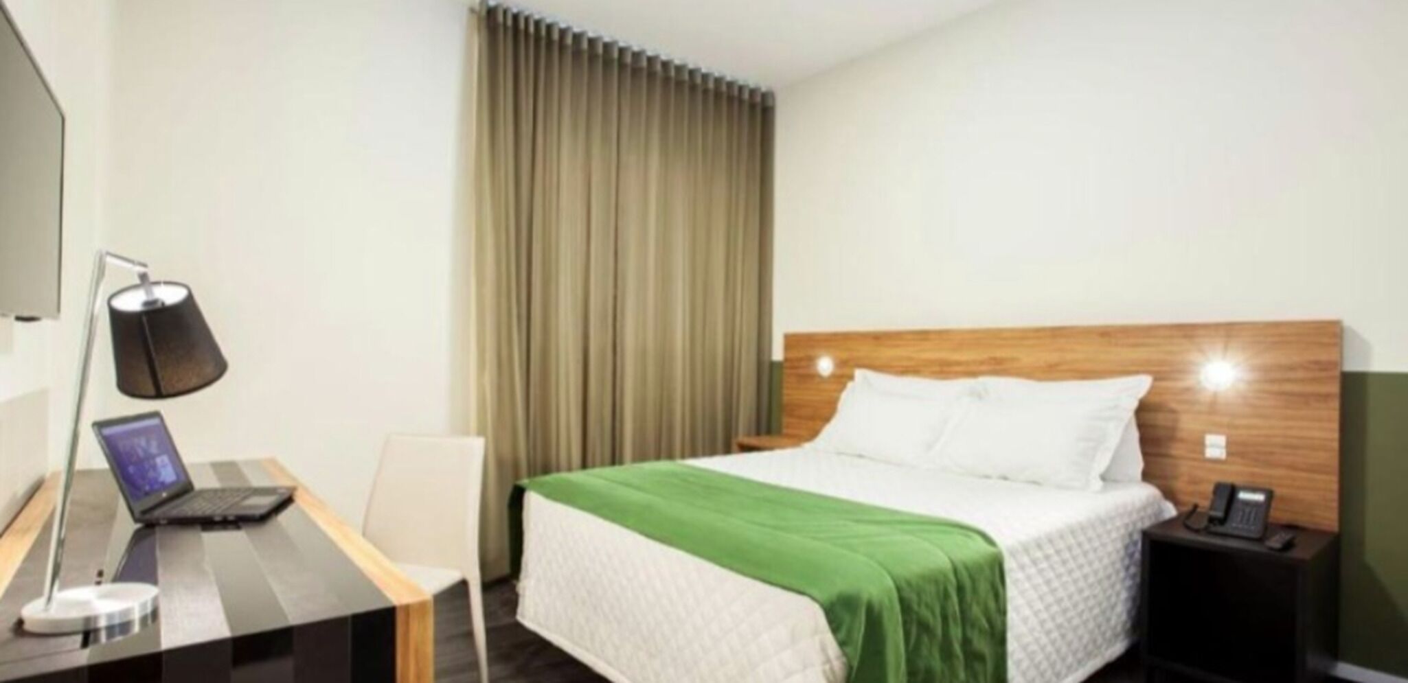 Una habitación de hotel con cama, escritorio, computadora portátil, lámpara, teléfono y cortina.