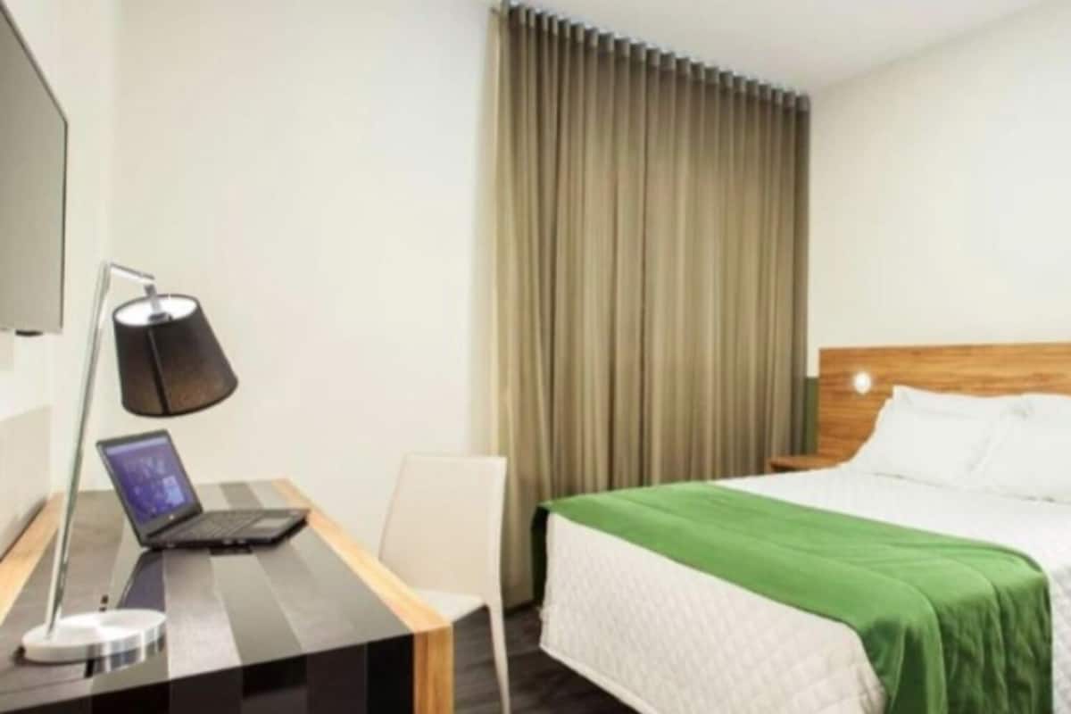 Une chambre d’hôtel avec un lit, un bureau avec un ordinateur portable, une lampe, un téléphone et un rideau.