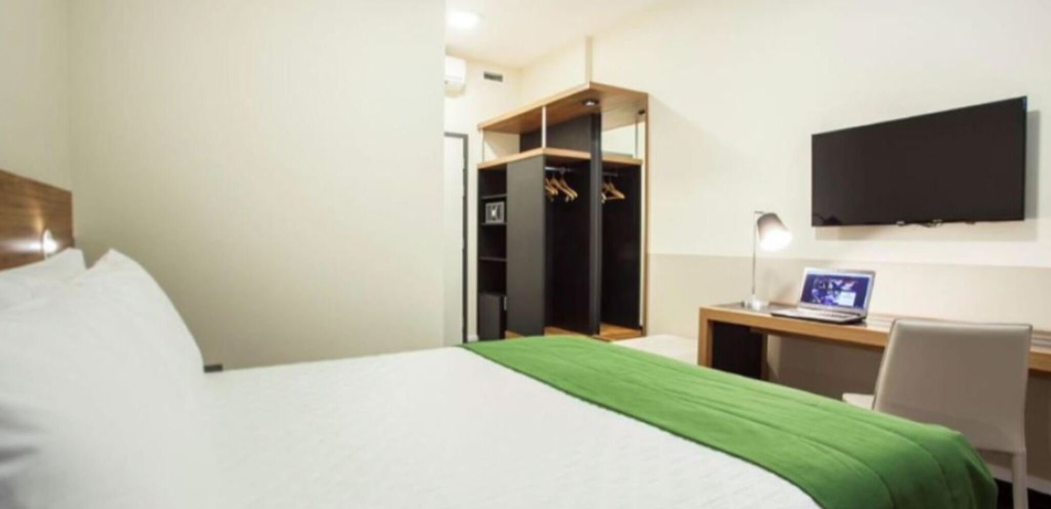 Habitación de hotel con cama, escritorio con una laptop, televisor montado en la pared y vestidor.