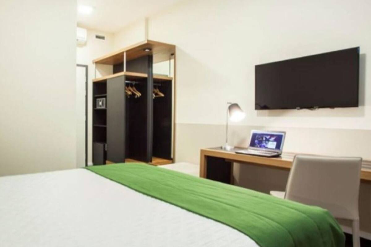 Une chambre d’hôtel comprenant un lit, un bureau avec un ordinateur portable, un téléviseur fixé au mur et un dressing.